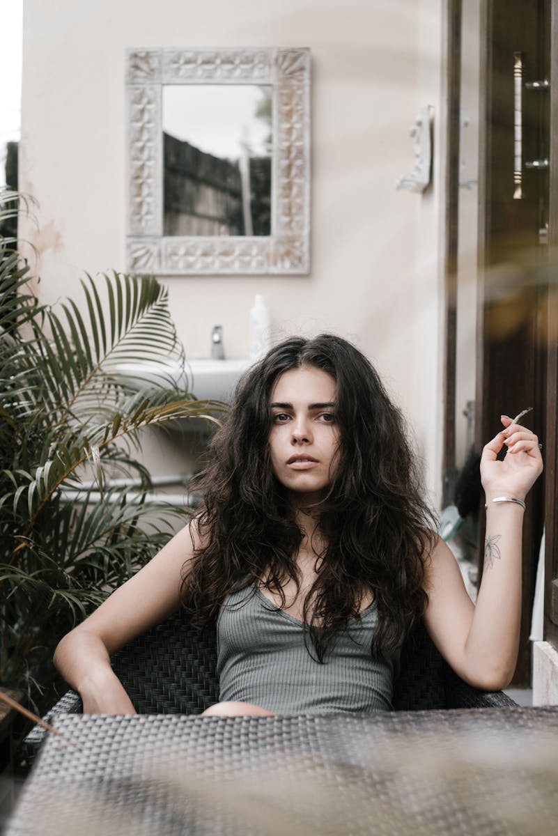 Content young female with long dark hair in summer top sitting at table on terrace with cigarette and looking at camera