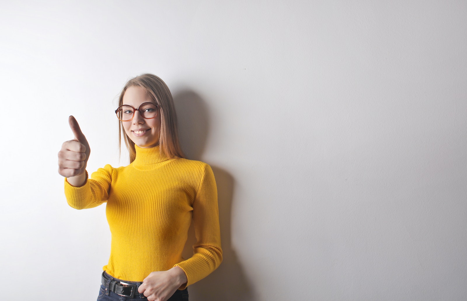 Photo of Woman in Yellow Turtleneck Sweater, Blue Denim Jeans, and Glasses Giving the Thumbs Up