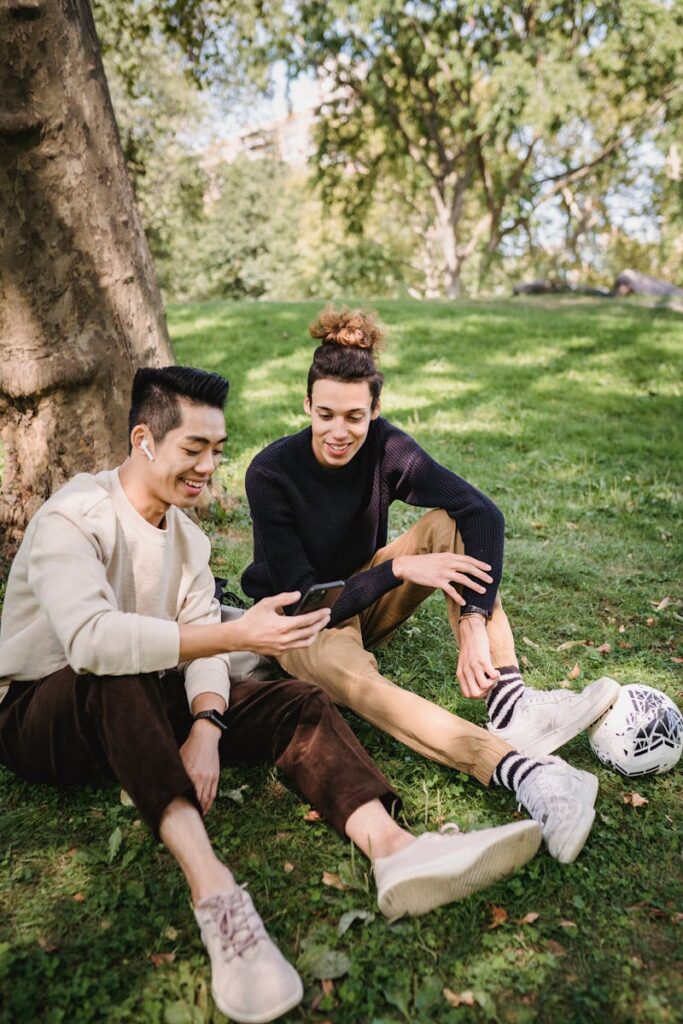 Smiling young ethnic males in casual clothes watching funny video on modern mobile phone while resting together on grass meadow in park